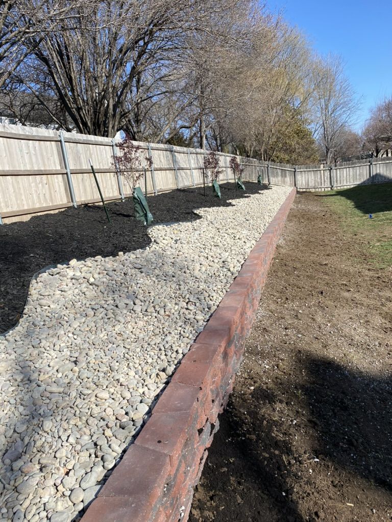 New trees planted in black mulch behind the river rock and retaining wall